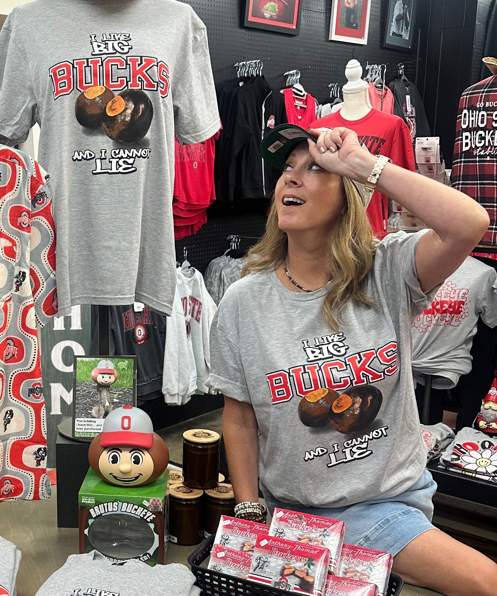 A woman wearing a gray Ohio Buckeyes t-shirt sits in a retail store next to a mannequin displaying a matching gray t-shirt with a cowboy hat. The gray t-shirt features 'The Ohio Big Buckies' and 'We're Making Lies' printed in red and orange.