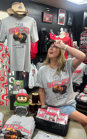 A woman wearing a gray Ohio Buckeyes t-shirt sits in a retail store next to a mannequin displaying a matching gray t-shirt with a cowboy hat. The gray t-shirt features 'The Ohio Big Buckies' and 'We're Making Lies' printed in red and orange.