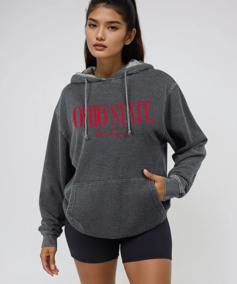 A woman modeling a dark gray heathered hoodie with 'OHIO STATE' printed in red across the chest, paired with black bike shorts, against a light gray studio background.