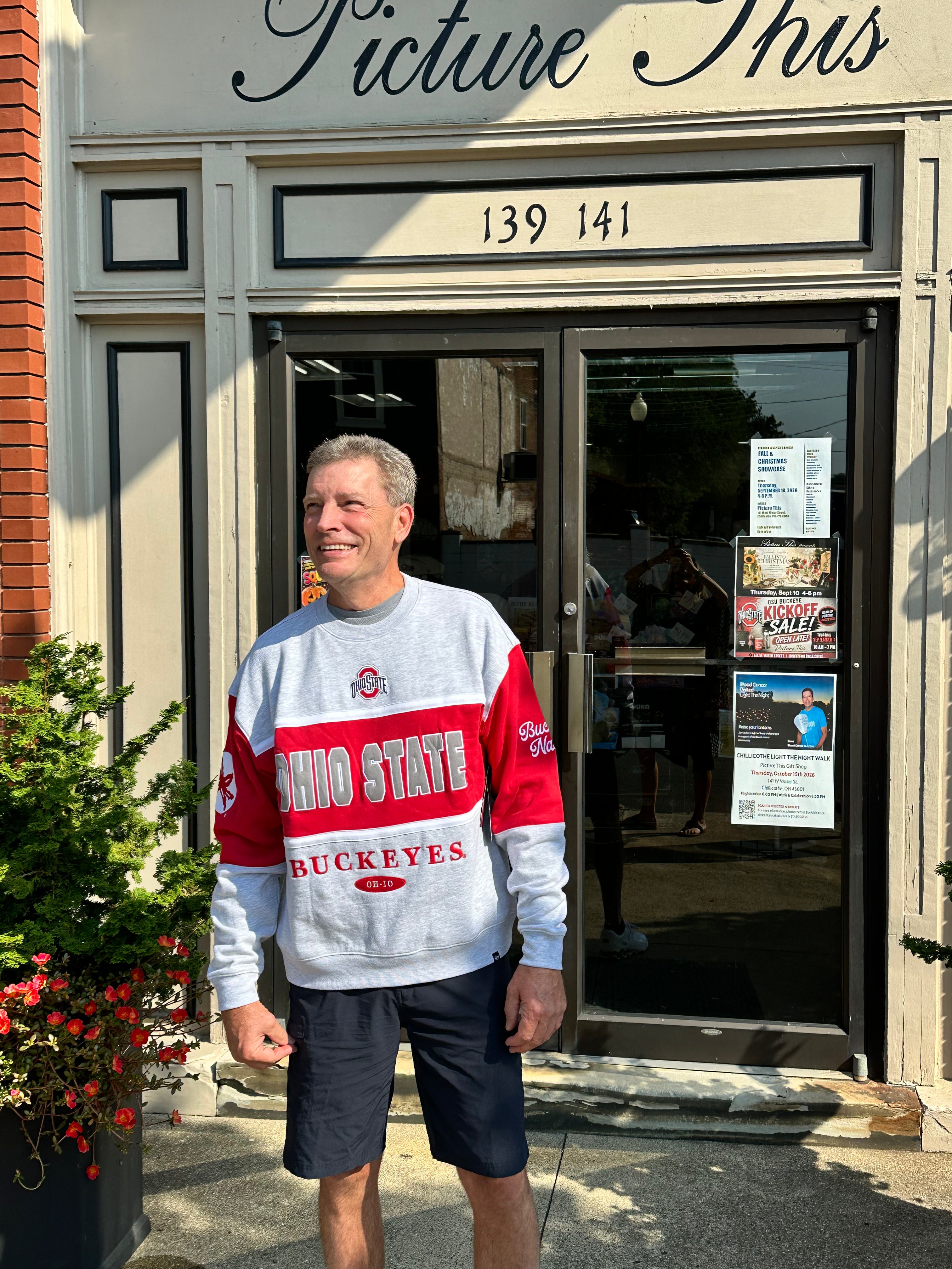 A man wearing a gray and red Ohio State Buckeyes sweatshirt and dark shorts stands in front of a storefront with 'Picture This' written above the door.