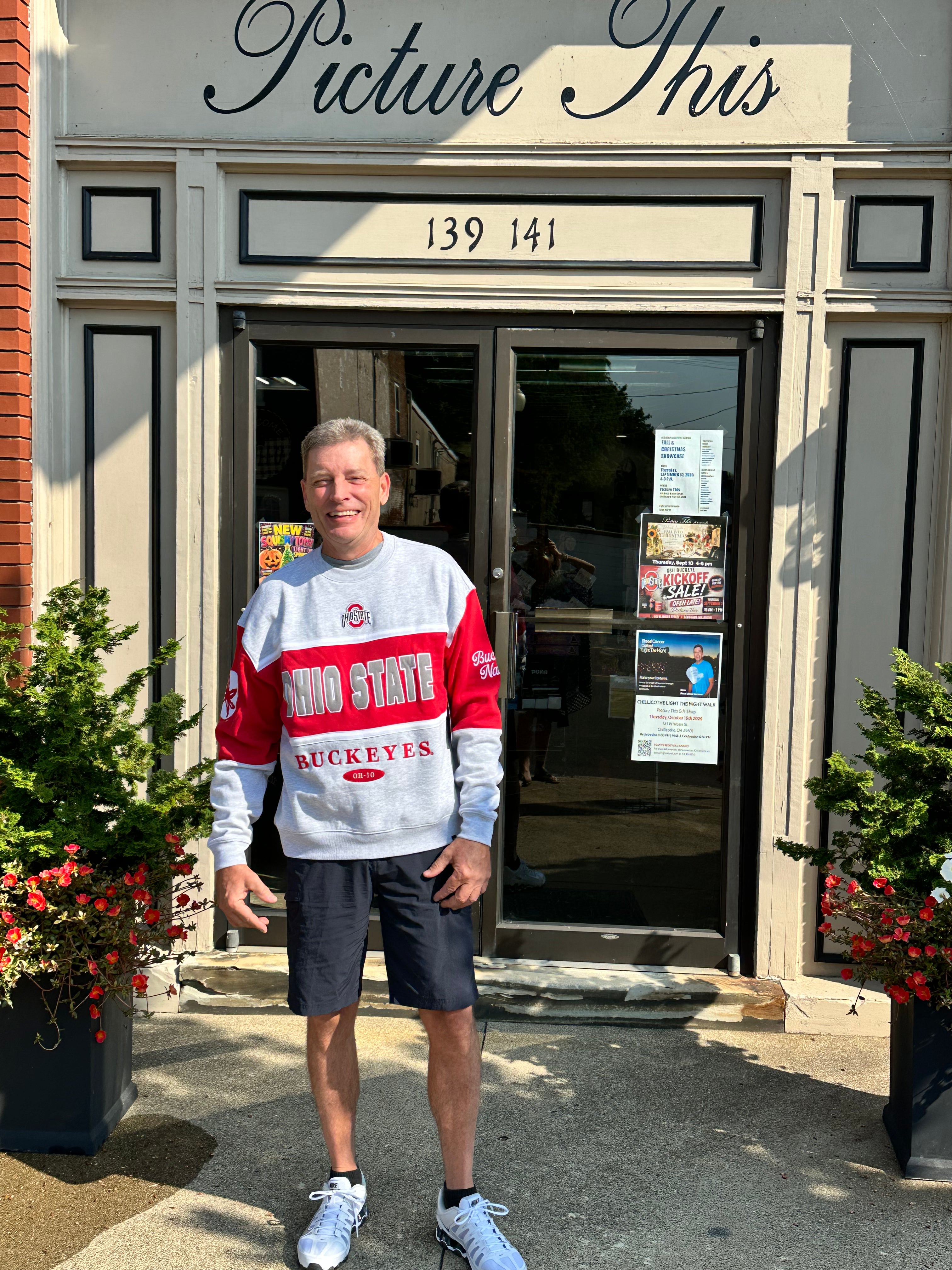 A man stands in front of a storefront wearing a gray and red Ohio State Buckeyes sweatshirt, dark shorts, and white sneakers. The shop has a sign reading 'Picture This' above the entrance.