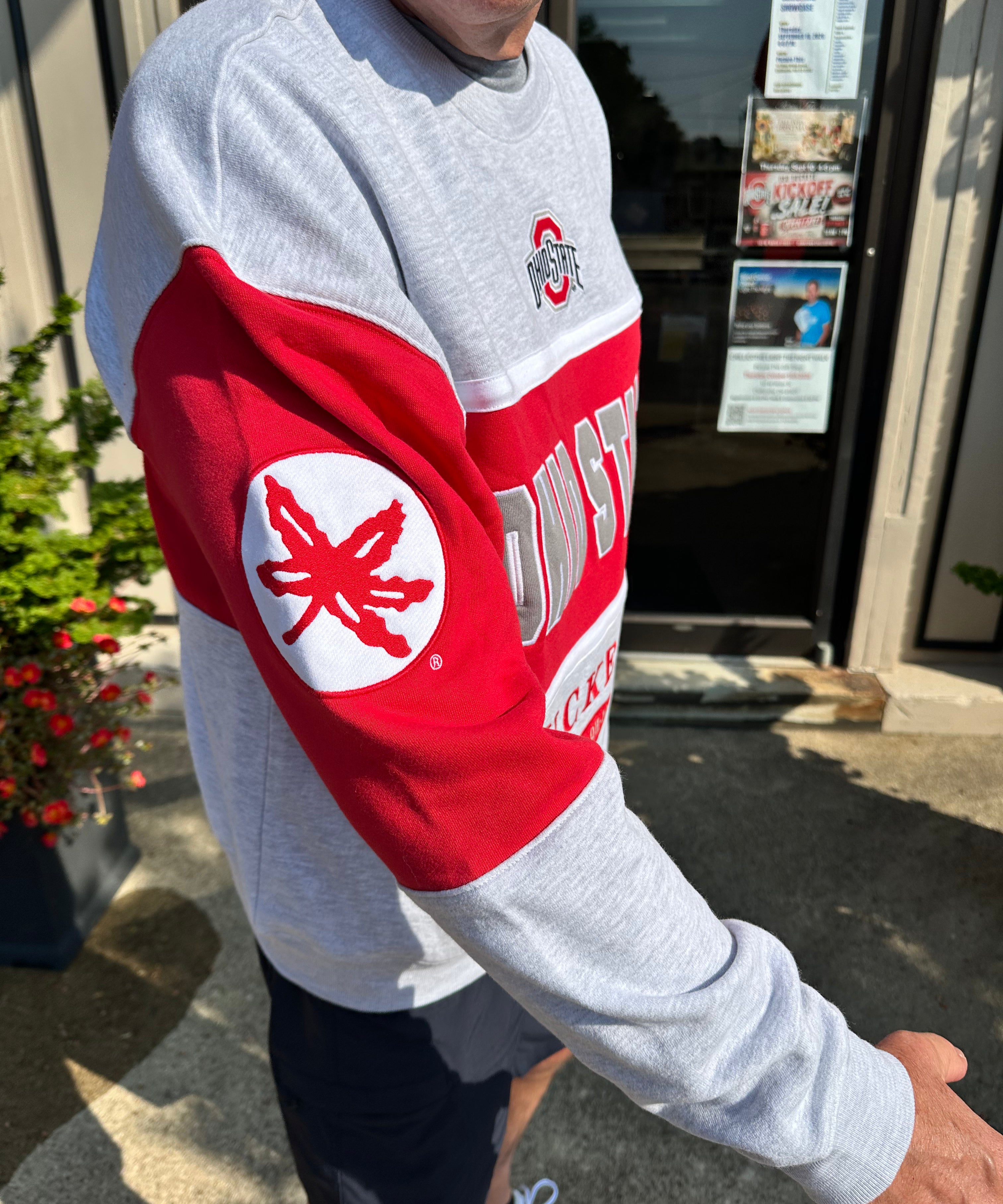 A person wearing a heather gray and red Ohio State sweatshirt, standing outdoors in front of a building entrance. The sweatshirt features a large red sleeve with a white circular patch containing a red Ohio State buckeye leaf, and 'OHIO STATE' printed across the upper arm.