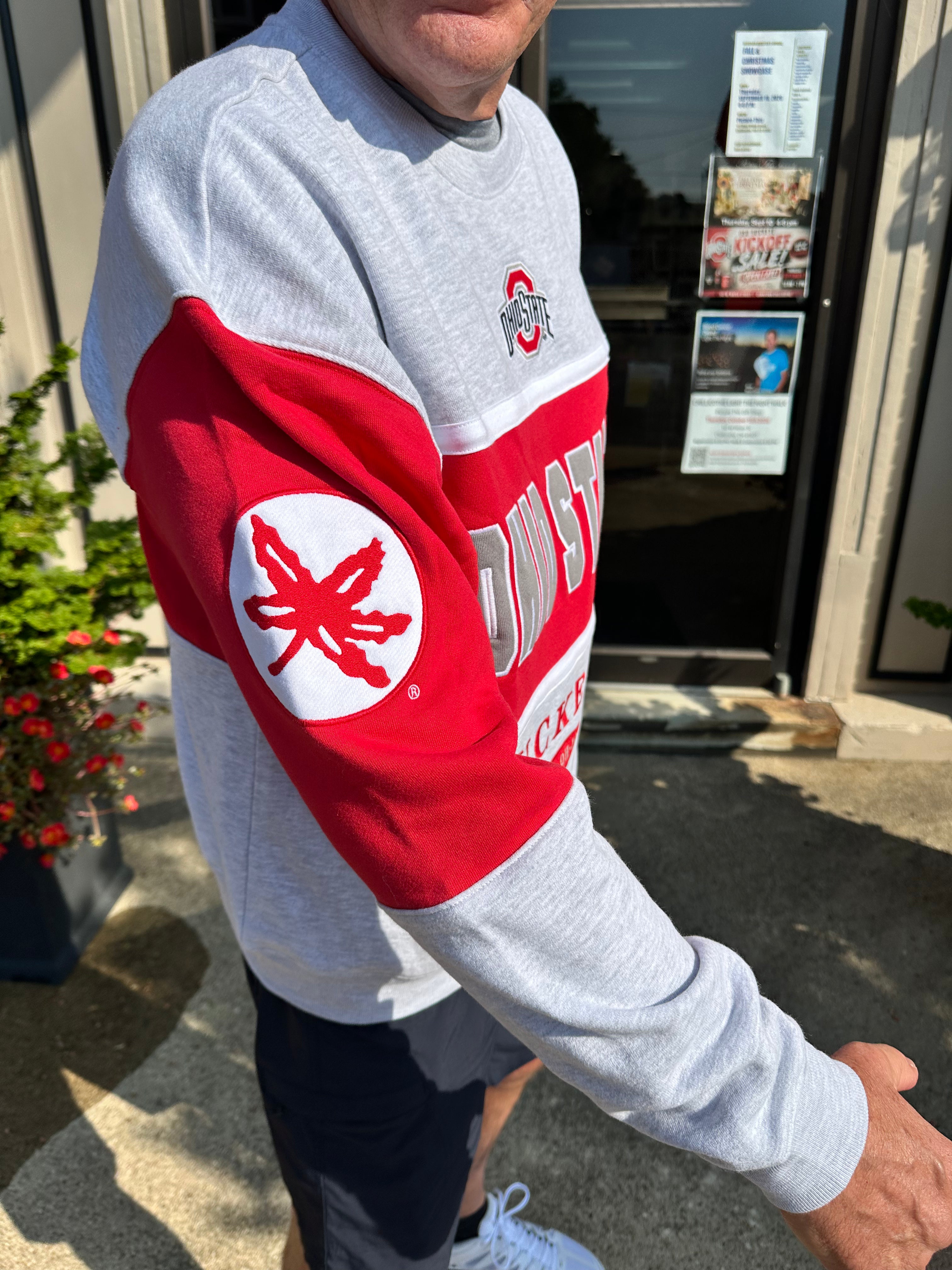 A person wearing a heather gray and red Ohio State sweatshirt, standing outdoors in front of a building entrance. The sweatshirt features a large red sleeve with a white circular patch containing a red Ohio State buckeye leaf, and 'OHIO STATE' printed across the upper arm.