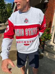 A man wearing a heather gray and red Ohio State Buckeyes sweatshirt, paired with dark gray shorts, standing outdoors on a sidewalk in front of a brick building.