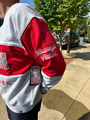 A person outdoors wearing a red and gray sweatshirt with 'Buckeye Nation' embroidered on the sleeve. The sweatshirt has a large gray block letter on the right shoulder and part of a red 'YES' on the left chest.