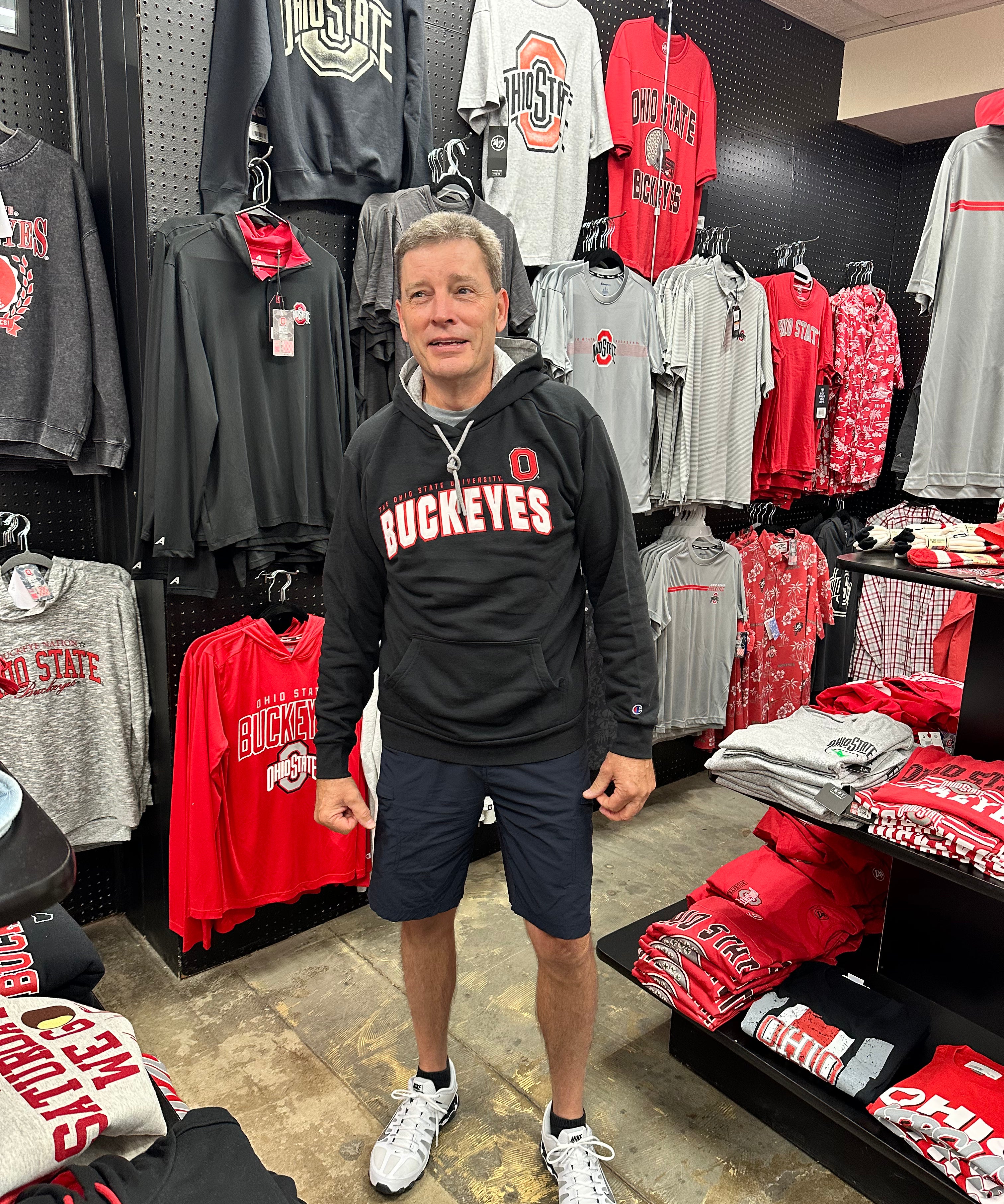 A man wearing a black Ohio State Buckeyes hoodie and navy shorts standing inside a retail store surrounded by Ohio State Buckeyes merchandise.