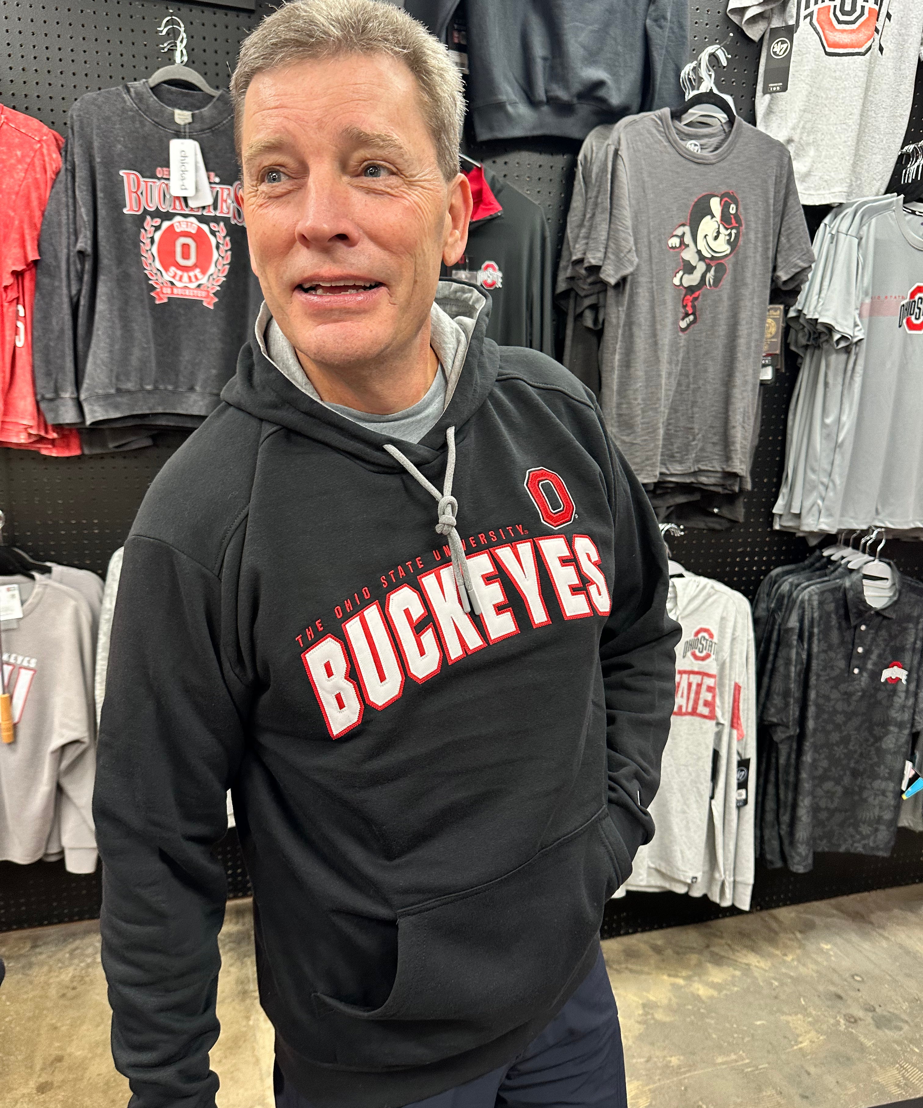 A man wearing a black Ohio State Buckeyes pullover hoodie stands in a retail store, with various Ohio State themed t-shirts hanging on the pegboard behind him.
