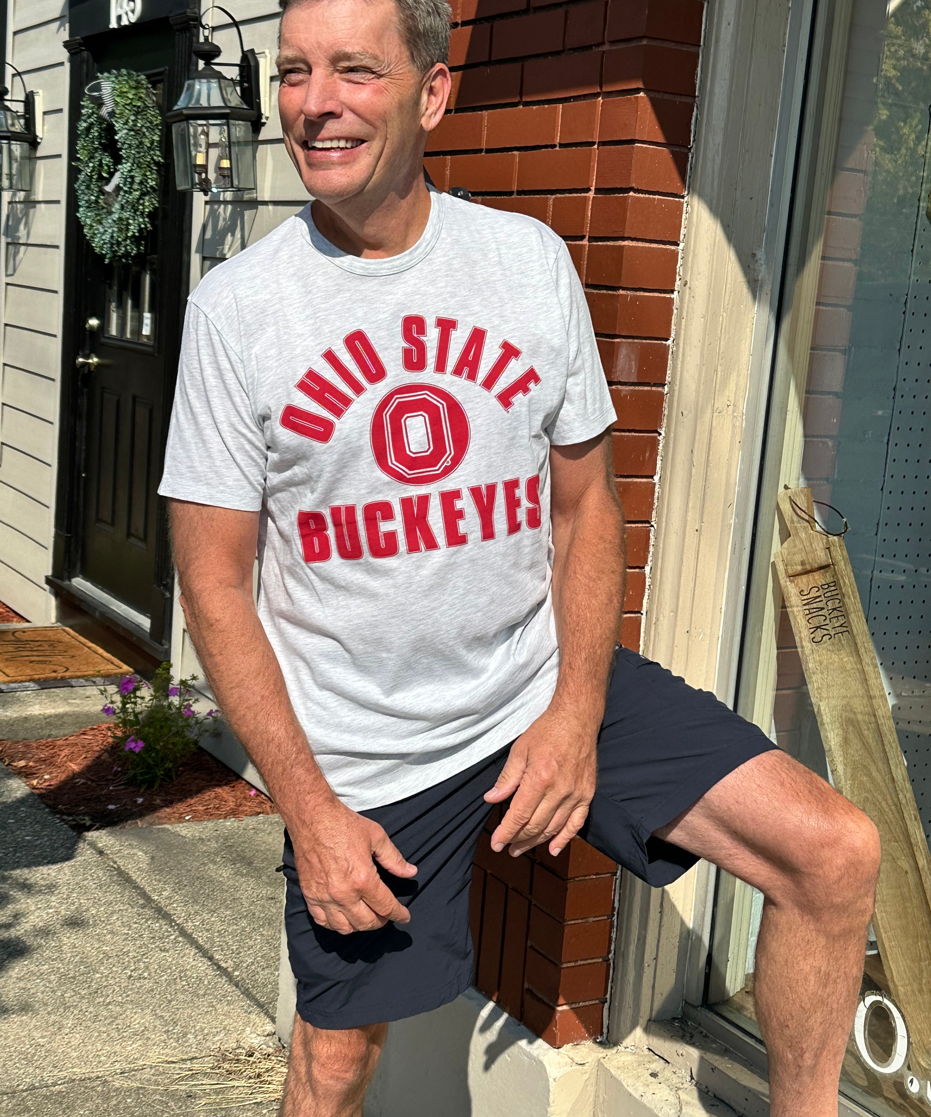 A smiling man wearing a light gray heather t-shirt with a red 'OHIO STATE BUCKEYES' graphic and dark navy shorts, posing outdoors against a brick and house facade.