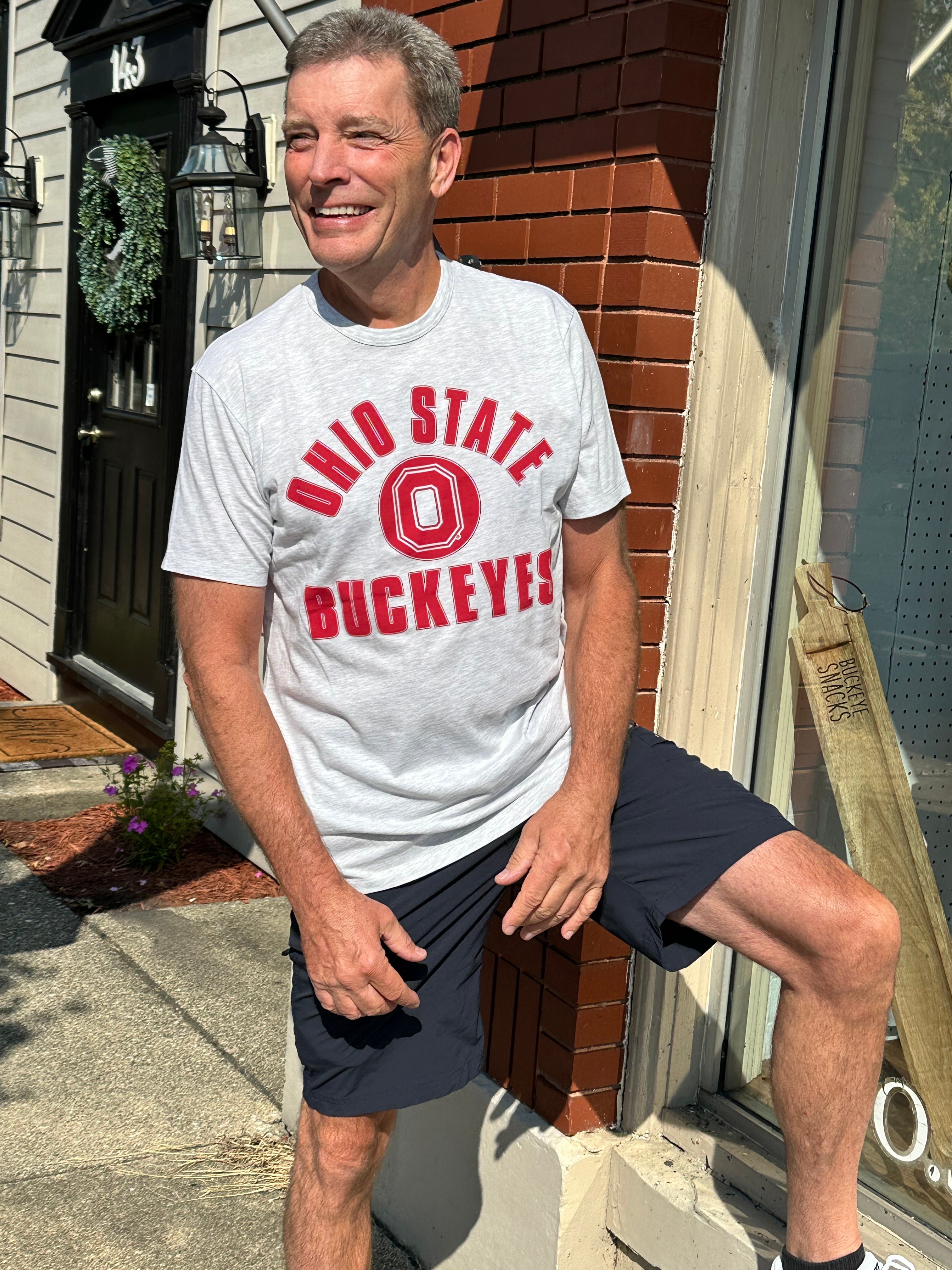 A smiling man wearing a light gray heather t-shirt with a red 'OHIO STATE BUCKEYES' graphic and dark navy shorts, posing outdoors against a brick and house facade.