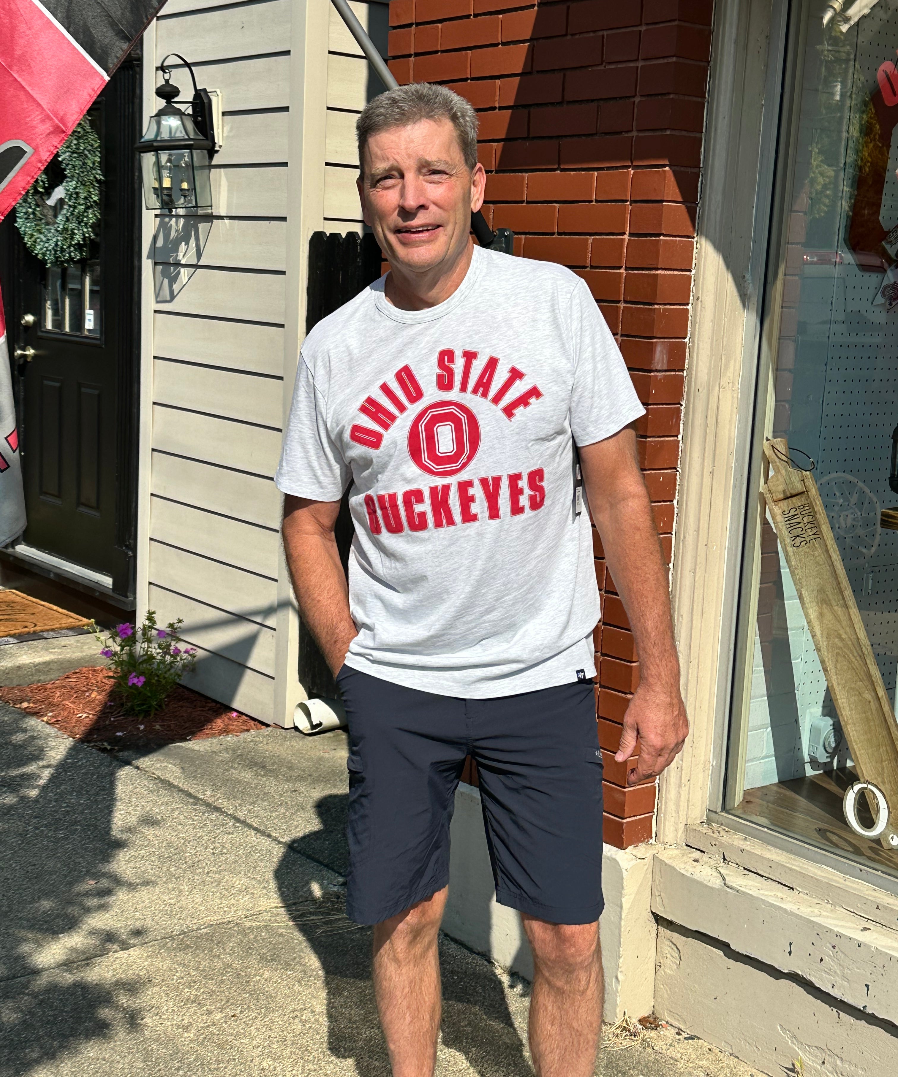 A man standing outdoors on a sidewalk in front of a brick and siding building, wearing a light gray t-shirt with red 'Ohio State Buckeyes' lettering and dark navy shorts.