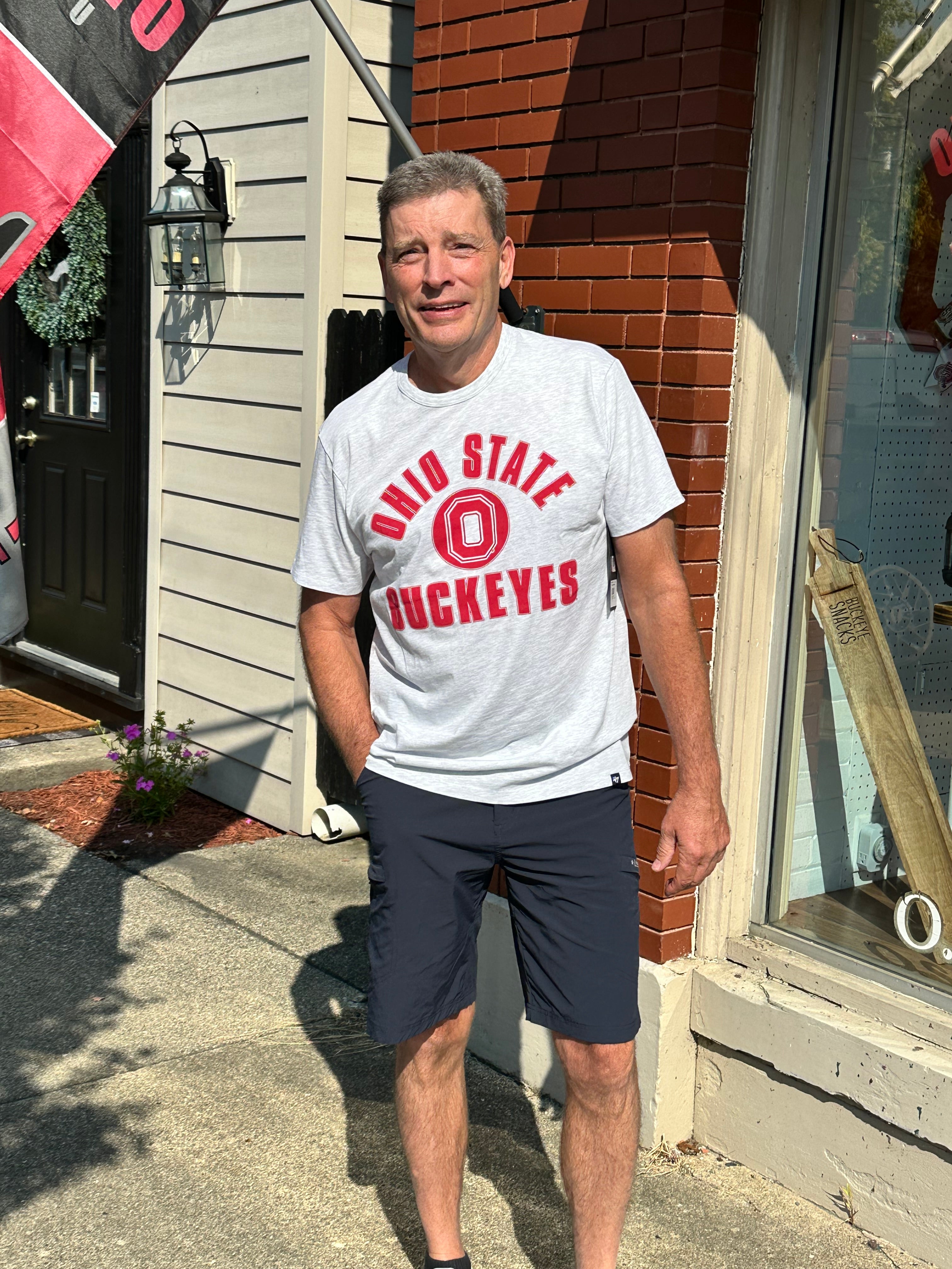 A man standing outdoors on a sidewalk in front of a brick and siding building, wearing a light gray t-shirt with red 'Ohio State Buckeyes' lettering and dark navy shorts.