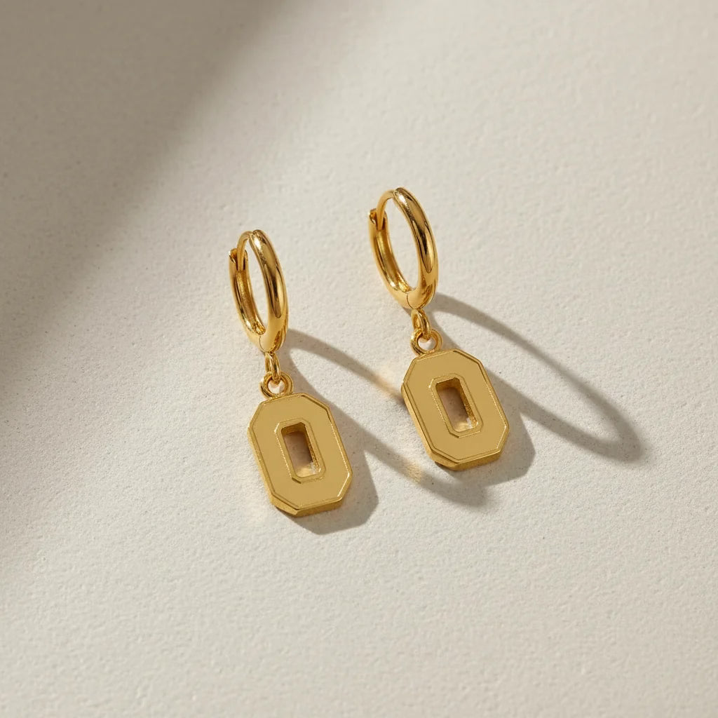 A pair of gold hoop earrings with flat, geometric rectangular drop charms featuring a cutout center, displayed on a textured off-white surface with soft shadows.