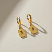 A pair of gold hoop earrings with flat, geometric rectangular drop charms featuring a cutout center, displayed on a textured off-white surface with soft shadows.