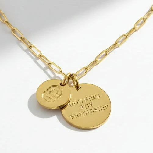 A gold paperclip chain necklace with two interlocking round gold pendants, one engraved with a geometric logo and the other with the engraved text 'LOYALTY FRIENDSHIP'.
