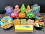 A collection of various novelty toys and squishy items arranged on a dark surface against a black background. The assortment includes a yellow butter bar labeled '4oz BUTTER', several colorful glittery squishies in pink, green, and orange, packaged plastic toys, and a 'nice CLUBE' box.