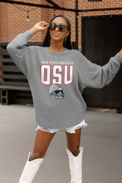 Woman wearing a gray sweatshirt with 'OSU' and a football helmet graphic, standing in front of a brick wall.