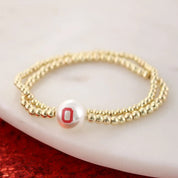 A gold beaded double-strand bracelet featuring a white pearl charm with a red letter 'O' emblem, resting on a white marble surface with a red glittery surface in the background.
