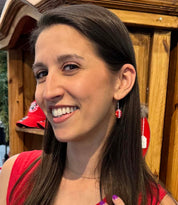 A smiling woman with long dark brown hair wearing a red top and a small red dangling earring with a white letter 'M' and a red heart, standing in front of a wooden display cabinet.